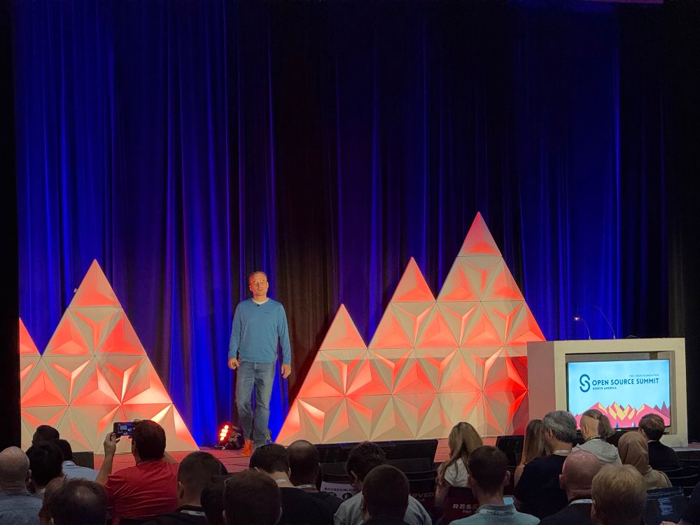A speaker stands on stage at the Open Source Summit North America, wearing a blue long-sleeve shirt and jeans. The stage features a red and white geometric mountain design in front of dark blue curtains. Audience members are seated and some are taking photos or using laptops. The event branding is displayed on a podium to the right.