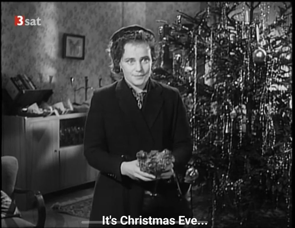 Maria Schell in overcoat and hat, holding a teddy bear. Behind her is a Christmas tree decked with tinsel and candles, and a sideboard. Subtitled "it's Christmas Eve..."