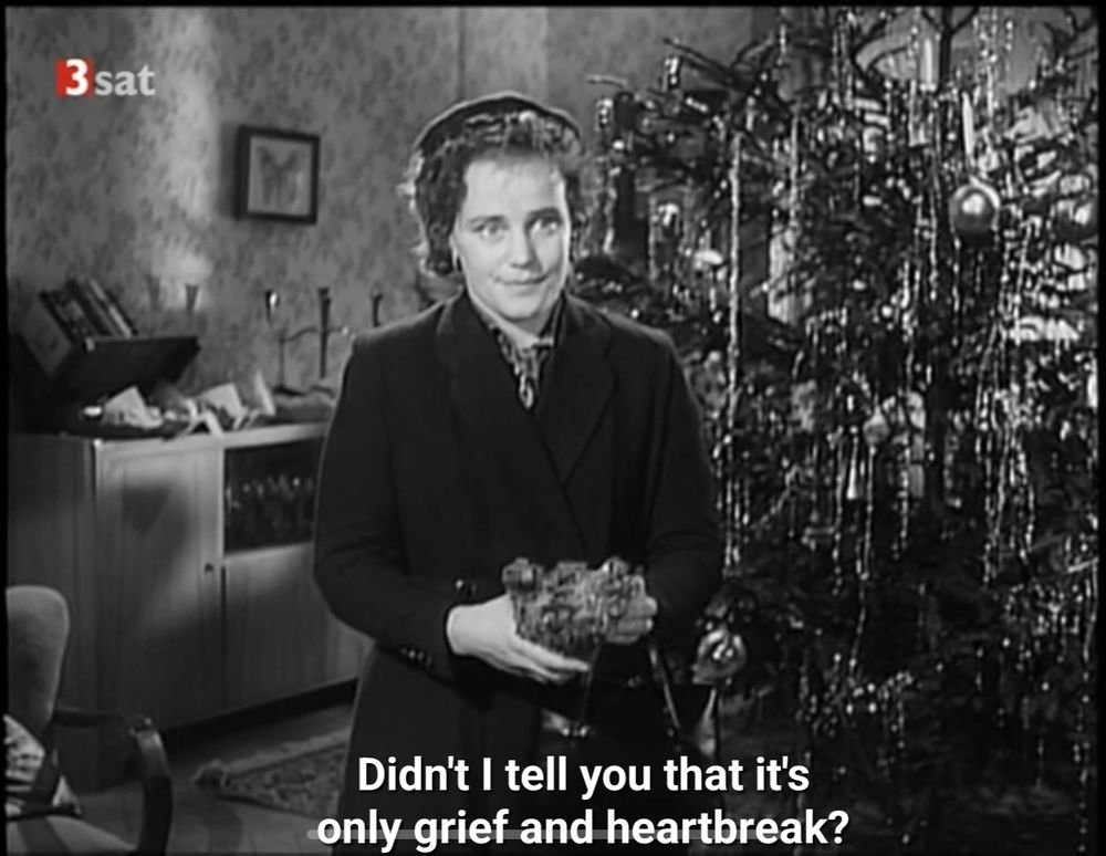 Maria Schell in overcoat and hat, holding a teddy bear. Behind her is a Christmas tree decked with tinsel and candles, and a sideboard. Subtitled "didn't I tell you that it's only grief and heartbreak?"