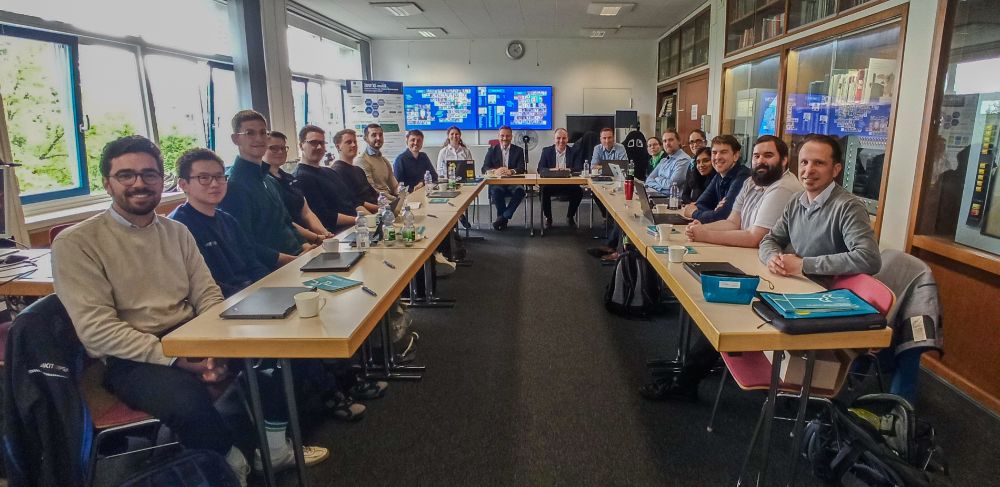 Gruppenfoto eines Meetings mit rund 20 Personen, die an zwei langen Tischen in U-Form sitzen. Die Teilnehmerinnen und Teilnehmer blicken lächelnd in die Kamera. Im Hintergrund sind Fenster, Poster und große Bildschirme mit Präsentationsfolien zu sehen. Die Atmosphäre wirkt professionell und kollegial.