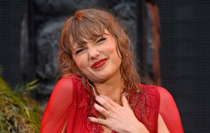 
This image shows a taylor swift performing on stage, wearing a vibrant red outfit with delicate details. She appear to be expressing strong emotion, smiling with their eyes closed and a hand over their chest. Her wavy, slightly wet-looking hair adds to the dramatic effect, and the background suggests a lively concert or performance atmosphere.