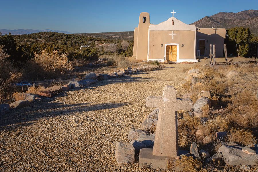 A brown stone cross in foreground with a southwestern adobe church with white painted trim in background. Clear blue skies, juniper and shrub landscape with low mountains. 