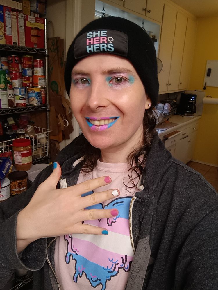 She hers beanie, blue pink white lipstick and eye shadow alongside nails and Pink trans flag heart shirt.