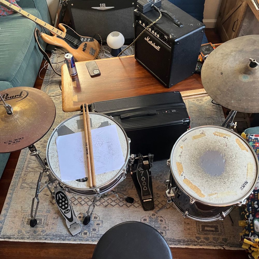 jank drumset with a suitcase as a bass drum. there is a bass, a redbull, and an amp behind.