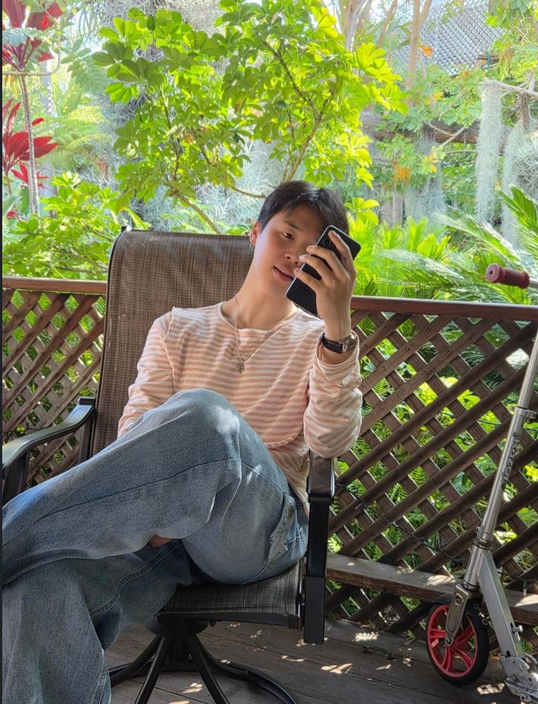jimin on @/uarmyhope insta dump, sitting on a chair outside, legs crossed and looking at his phone.