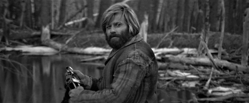 Capture d'écran du film Jeremiah Johnson de Sydney Pollack (1972) :  Jeremiah Johnson (Robert Redford), un trappeur chevelu et barbu vêtu d'un manteau regarde par-dessus son épaule gauche, tenant un piège à castor près d'une rivière dont la rive est partiellement enneigée.
