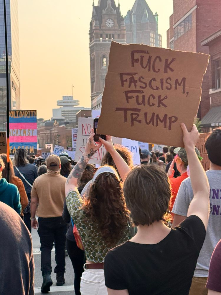 A photo of a group of protesters marching through Milwaukee, from behind. A prominent sign reads “Fuck Fascism, Fuck Trump.” Another reads “Solidarity Against Fascism” across a background matching the colors of the transgender pride flag.