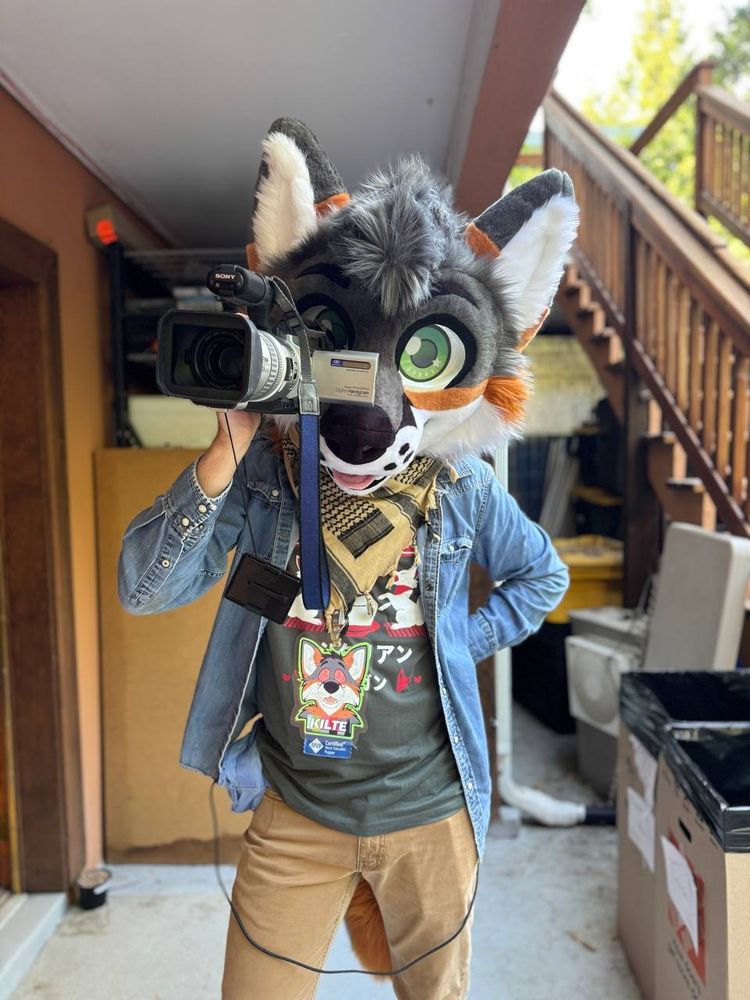 Me looking at the cmera while holding a sony miniDV vx2000 camcorder up to my fursuit head. I'm wearing chinos, a gray graphic tee, blue denim jacket, and a tan keffiyeh around my neck. A con badge is clipped to the keffiyeh