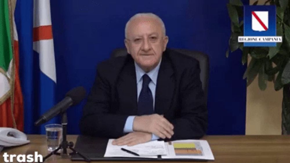 a man in a suit and tie sits at a desk in front of a microphone with a sign that says trash