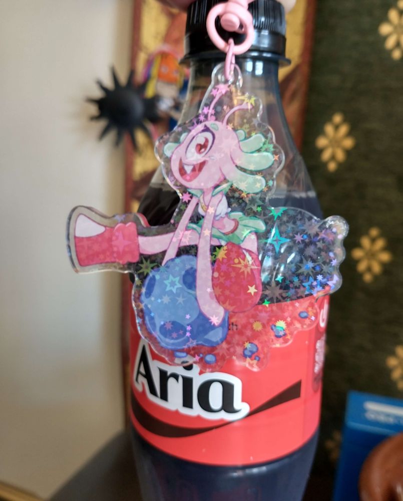 A keychain of Aria, hovering infront of a coke bottle with "share a coke with Aria" written on it