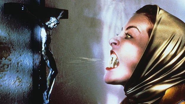 A still from the movie "The Lair of the White Worm" that depicts the antagonist, Lady Sylvia Marsh, manifesting fangs and spitting venom on a crucifix inside of Mercy Farm.
