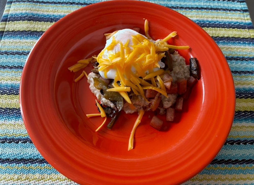 A red Fiesta bowl filled with savory steel cut oats topped with portobello mushrooms, spam, onion, bell pepper, a runny egg and cheddar cheese
