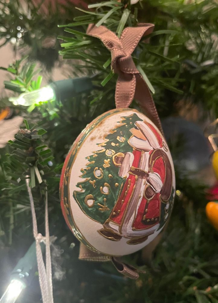 Ornament that is egg-shaped and illustrated primitive-style with Santa Claus in front of a festively decorated Christmas tree.