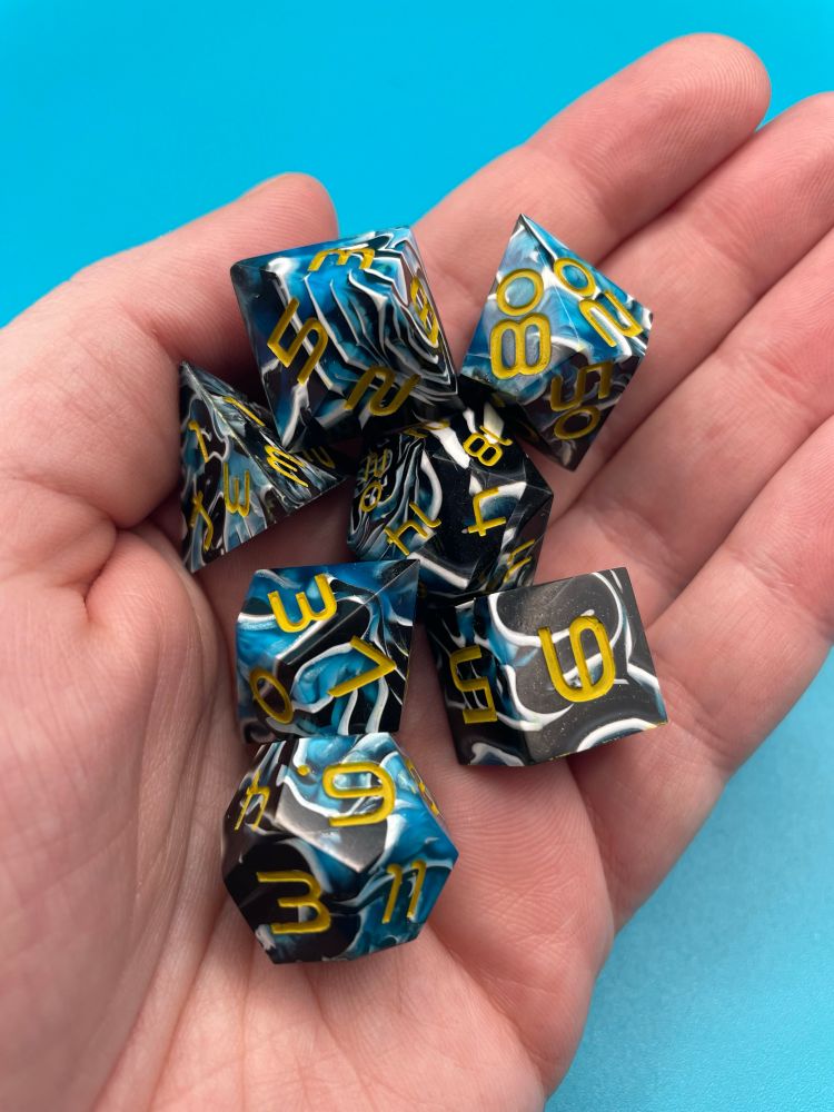 Photo of a set of ttrpg dice in a hand. The dice are swirls blue, black and white and have yellow numbers. 