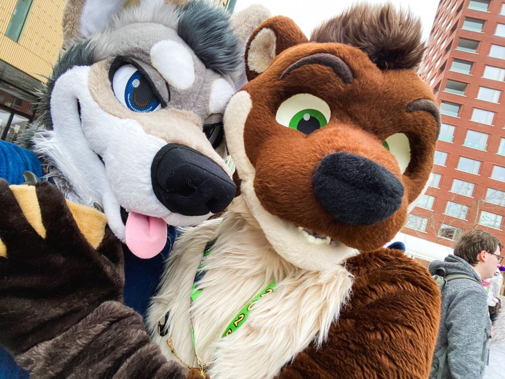 A close-up photo of two Fursuiters. On the left a white, light-brown Coyote Fursuiter with blue eyes named Mahkun and on the right a cream-and-brown colored Otter Fursuiter with green eyes named Blackymoon. They are both hugging each other from the side, looking at the camera that Mahkun himself is holding to take a selfie. Blackymoon has a webbed handpaw on Mahkuns Chest. Behind them are several people and Fursuiters as well as buildings visible.