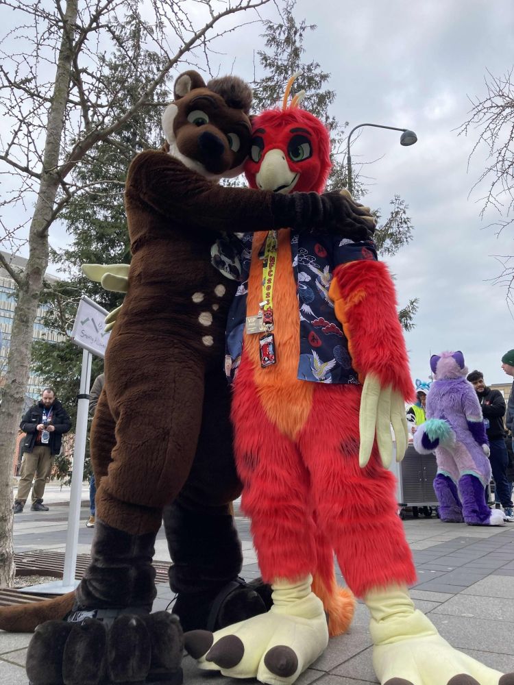 A big, cream-and-brown colored Otter Fursuiter named Blackymoon hugging a big, yellow-red-and-orange colored Phoenix Fursuiter named Phonixx from the side while both are looking at the viewer. Blackymoon has one arm around Phonixx's front shoulder and the other around his back while Phonixx has one of his wing-hands around Blackymoons waist. The view is slightly from down-below, giving both their footpaws a prominent size and making them both appear huge. They're standing outside a Convention Hotel on gray stone flooring with several other people and Fursuiters in the background.