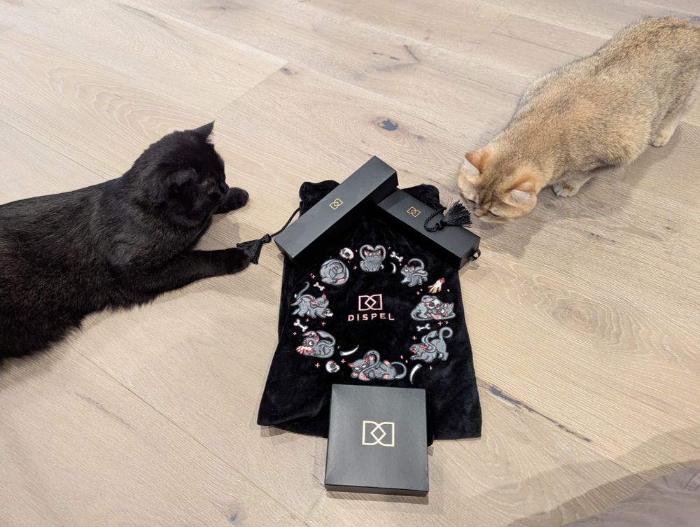 A tentacle kitten lucky bag from Dispel Dice is on the floor, covered with dice boxes. Grizzly, a black cat, is lying on the floor and watching as veteran QA feline, Uschi, appears to demonstrate proper sniffing technique of new Dispel Dice Merch