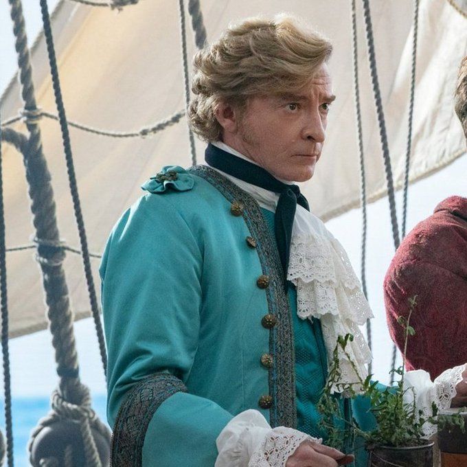 Stede Bonnet in teal jacket with buttons, black cravat, and gloriously tousled hair, facing right, with hand on decorative plant, in front of sail.