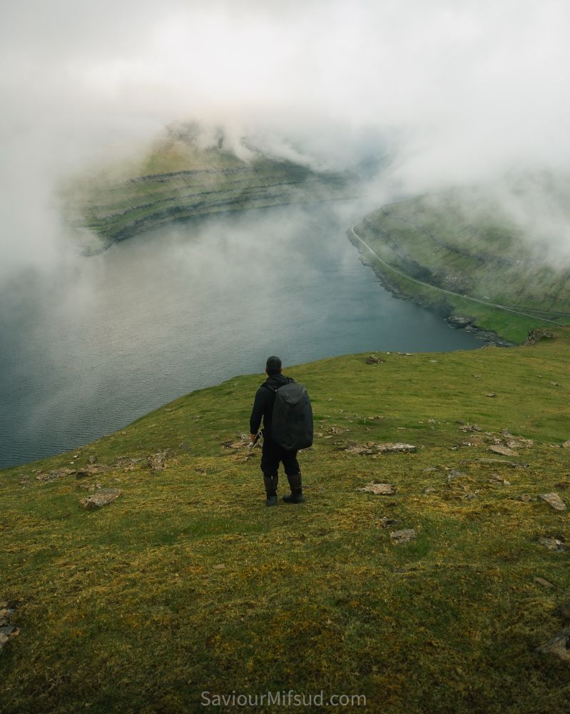 Man on top of mountain enjoying the view of Funningsfjørður fjord with some incredible weather conditions of low clouds covering the mountain peaks In the background.