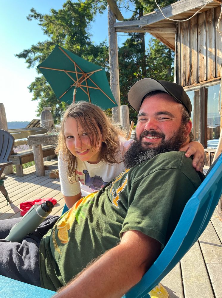 Graybeard son sits on a wooden deck in the Pacific Northwest with a black ballcap and a green T-shirt. He is smiling and looks high. My 10-year-old daughter leans over him smiling-ish to pose for pic, a green umbrella growing out of her head and blue sky behind.  