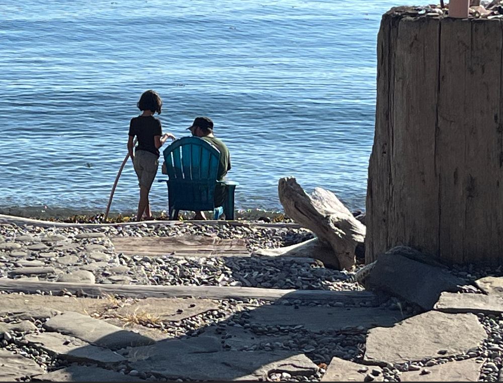 Jimmy from behind sitting in a chair down at the rocky shore in the San Juan islands. His skinny daughter stands silhouetted next to him with her feet in the water. 