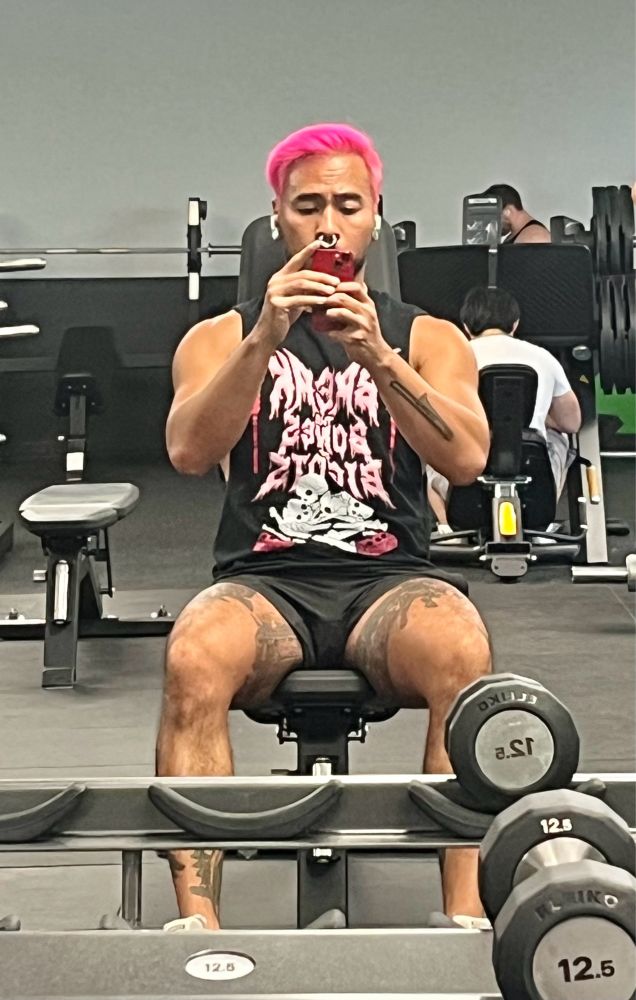 I’m an Asian guy with pink hair and a big nose ring, taking a mirror selfie sitting on a bench at the gym. I’m wearing a black and pink tank top and black shorts.