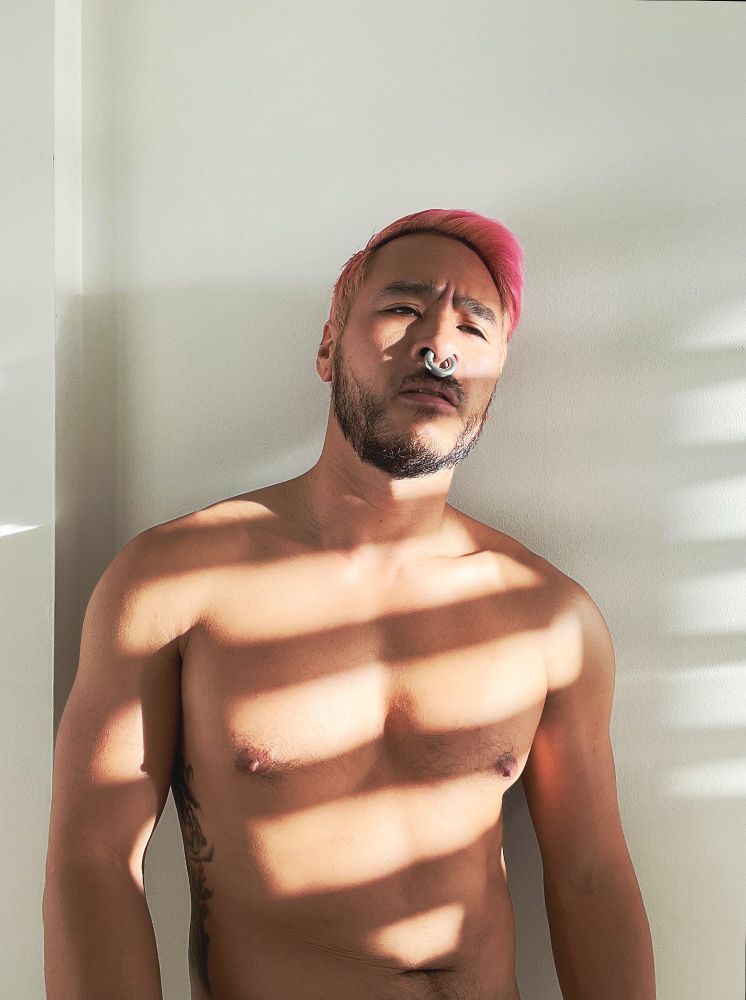I'm a pink haired Asian guy with a big nose ring, shirtless standing against a white wall with striped light across my body from window blinds.