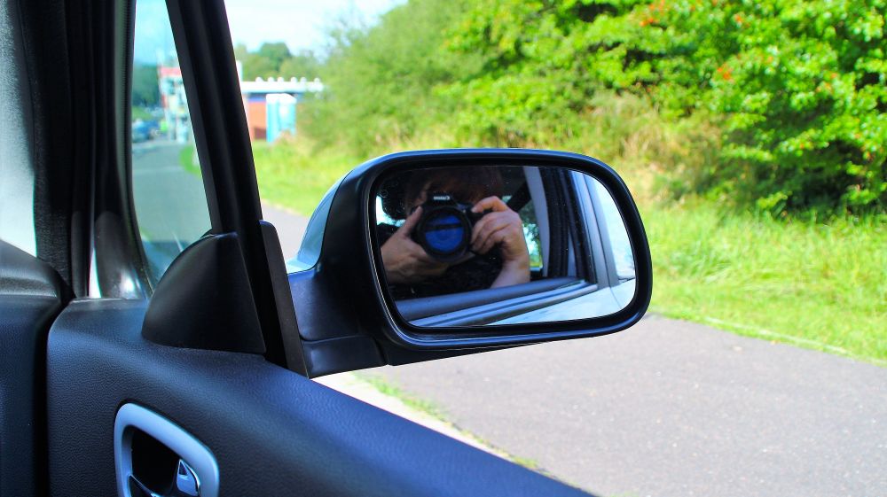 Ein Seitenrückspiegel eines Autos, im Spiegel erkennbar zwei Hände und ein Fotoapparat. Vor dem Auto verschwommen, die Fähre Landwehr am Nord-Ostsee-Kanal
