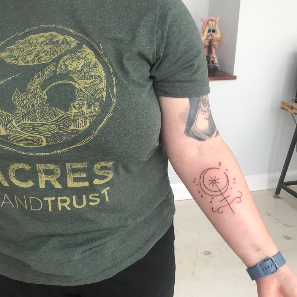 A feminine individual showing a fresh minimalist tattoo on her inner forearm. She is wearing a green t-shirt.