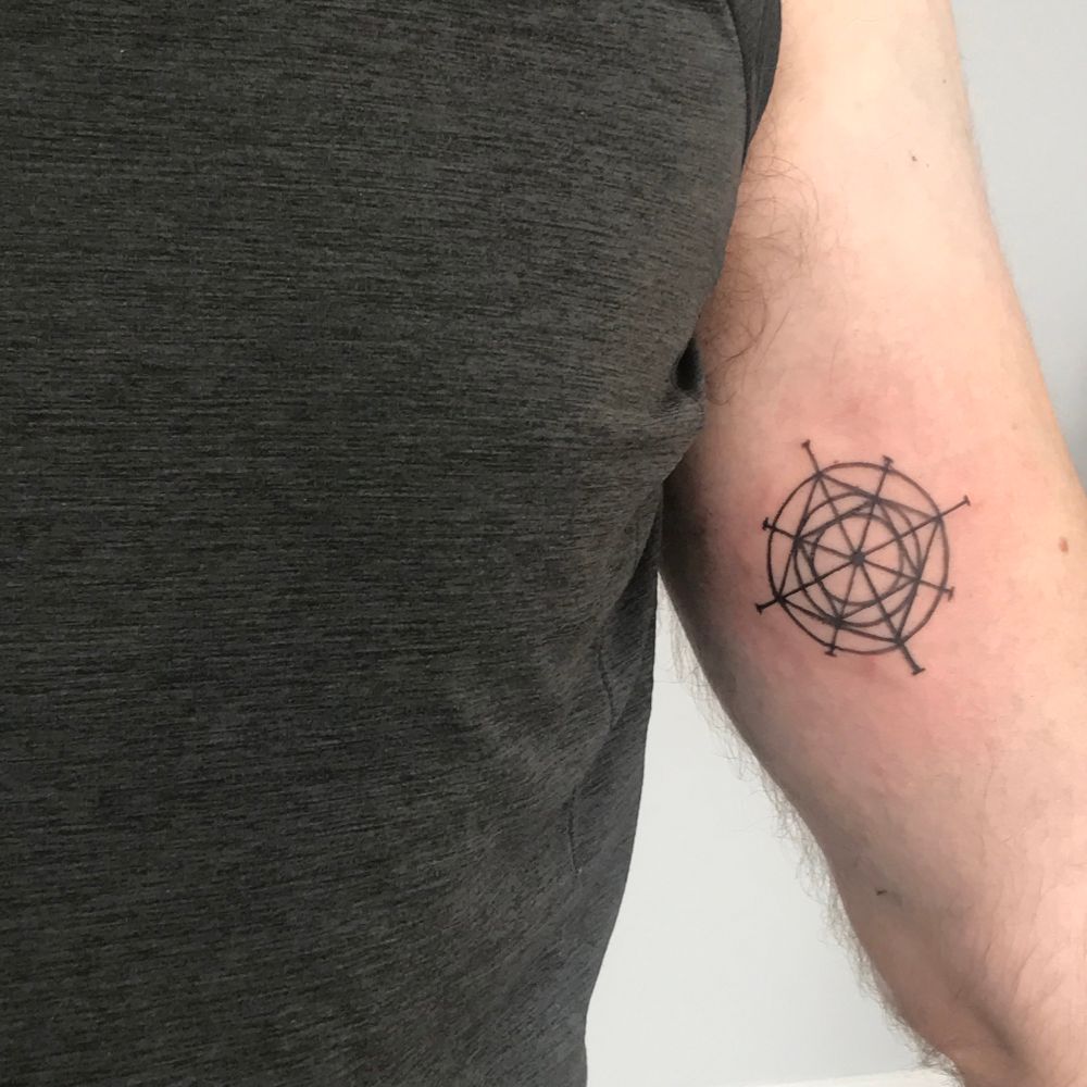 A photo of a masculine individual showing a fresh geometric tattoo on his inner bicep. He is wearing a grey shirt and is standing in front of an off-white wall.