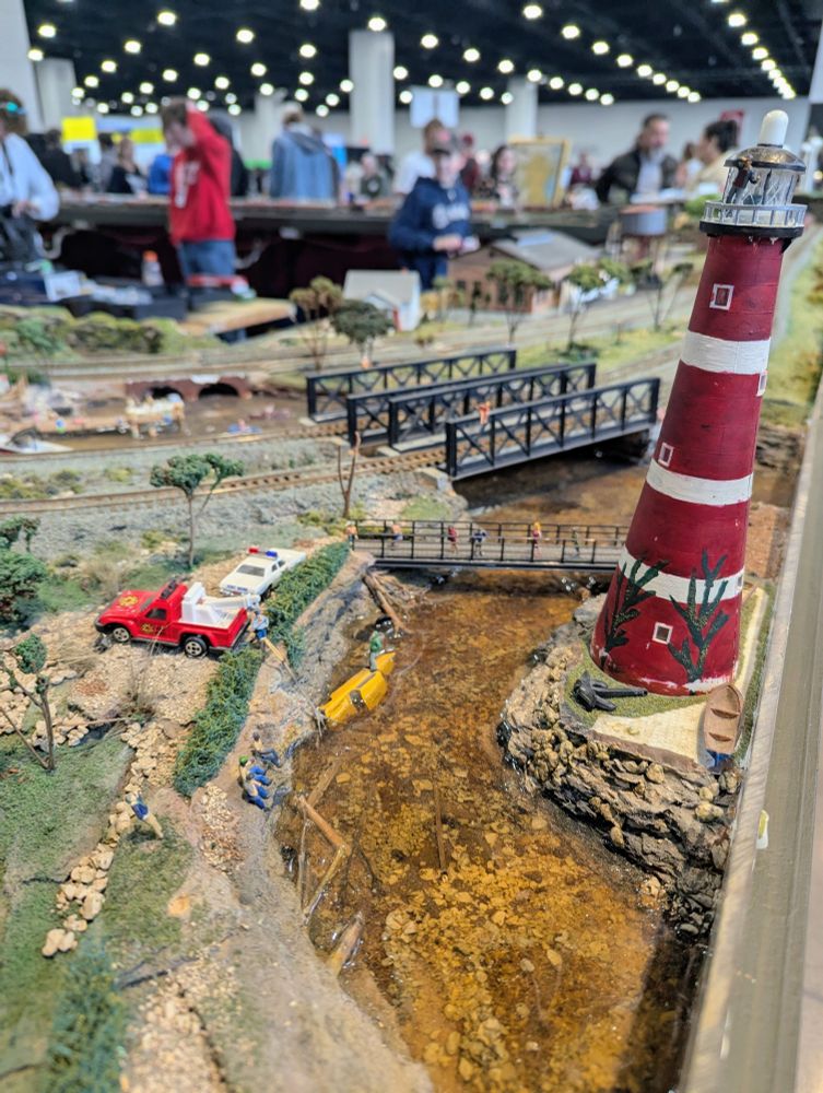 Model train layout with a lighthouse and a car that has fallen into the river. The huge conference hall space is visible out of focus in the background. 