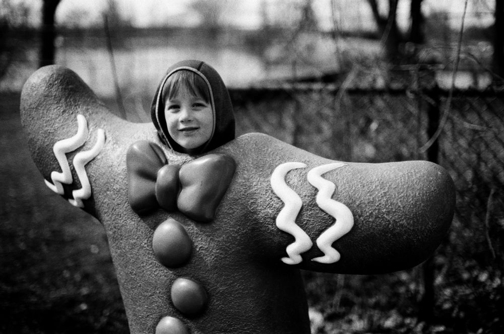 A black and white photograph of a young boy's head poking up out of the top of a gingerbread man sculpture 