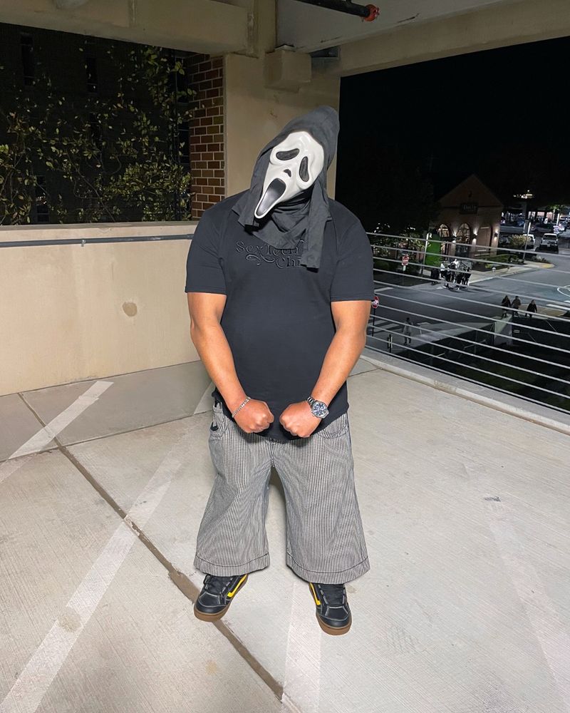 Reuben, a Black man flexing his muscles in a parking garage wearing a black t-shirt and Ghosteface mask. 