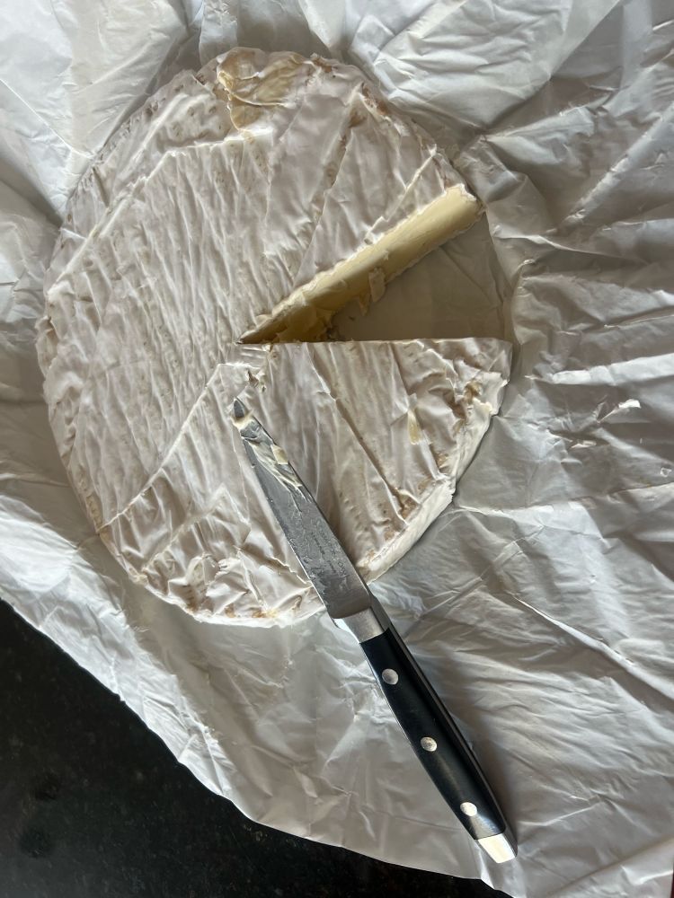 Photo of a wheel of Brie cheese with a slice cut out of it. Wheel is probably 8-10” diameter