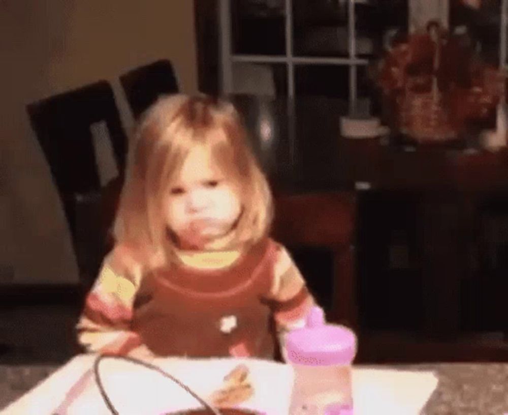 a little girl is sitting at a table with a plate of food and a pink bottle .