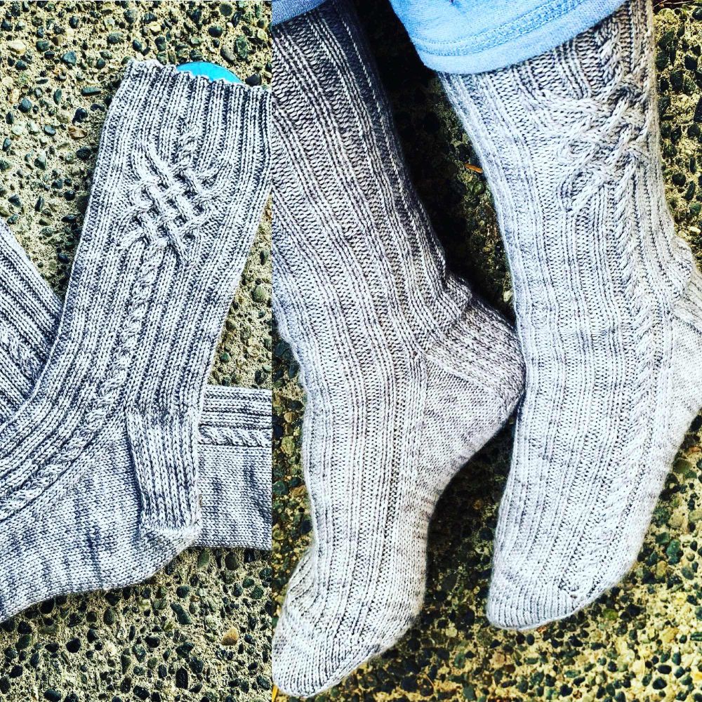 Split photo showing hand-knit silver gray cabled and ribbed women’s calf-length socks on sock blocker laying on pebbles ground on left and same socks being worn on feet on same background to right