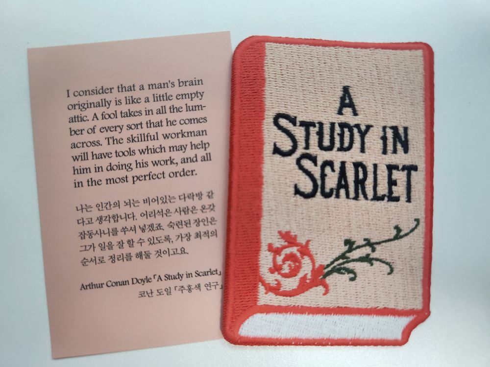 와팬 재질의 셜록 홈즈 시리즈의 첫 권인 a study in scarlet 책 모양 책갈피. 책은 주홍색이며 제목이 검은색 대문자로 쓰여 있다. 덤불 같은 것도 그려져 있다.