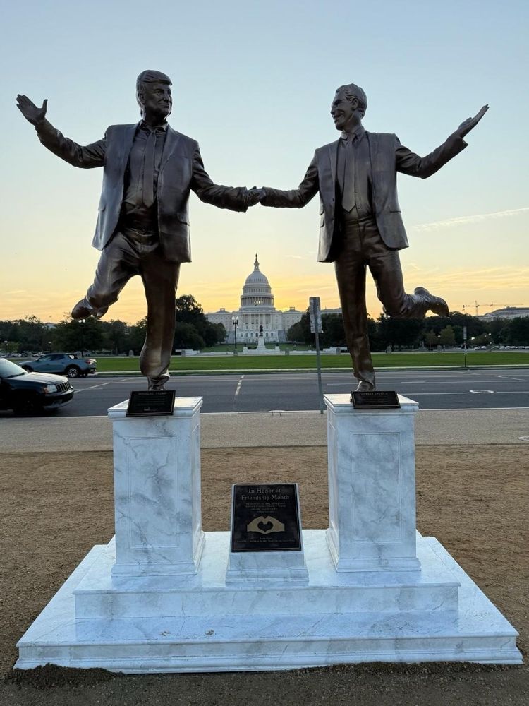 A statue with a white marble base and two podiums on which two male figures are smiling and holding hands in a stance that can only be described as frolicking