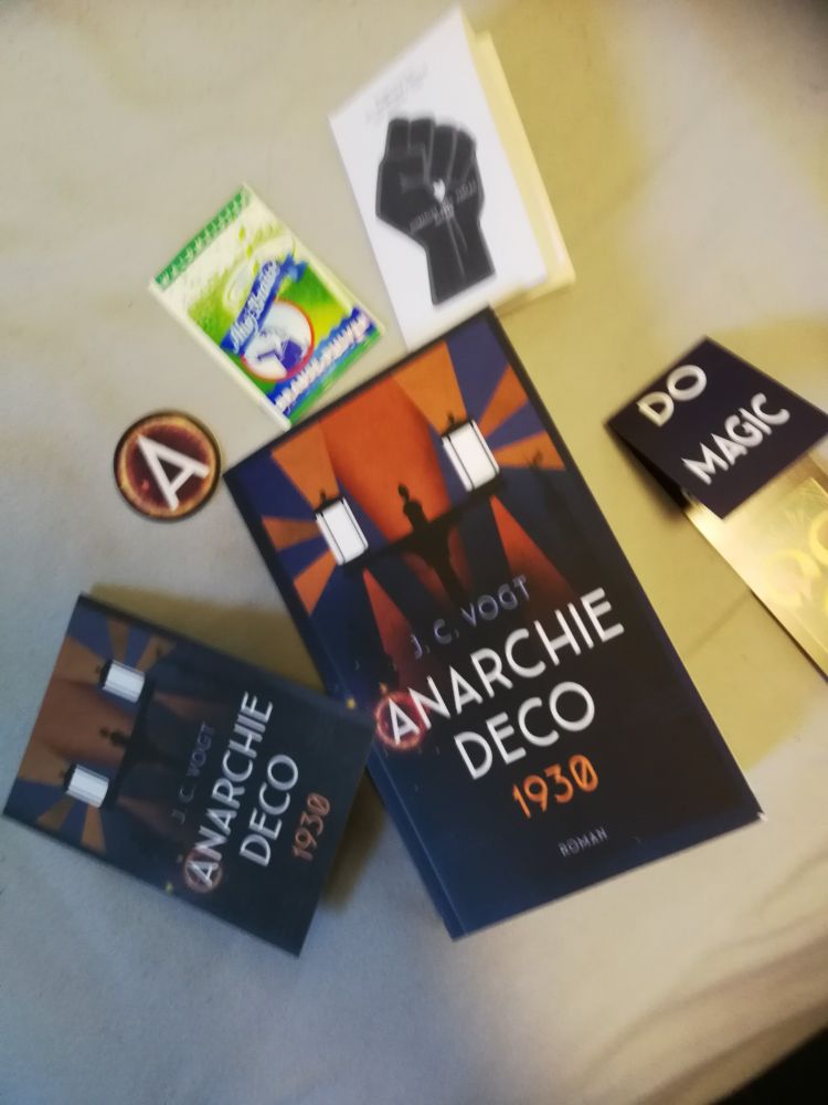Ein Exemplar von "Anarchie Deco 1930", umgeben von einer Postkarte mit dem Buchcover als Motiv, einem Aufkleber mit einem "A", einem Päckchen Ahoi Brause Waldmeister, einem "Flugblatt der Schwarzen Scharen" und einem Lesezeichen mit der Aufschrift "Do Magic". 