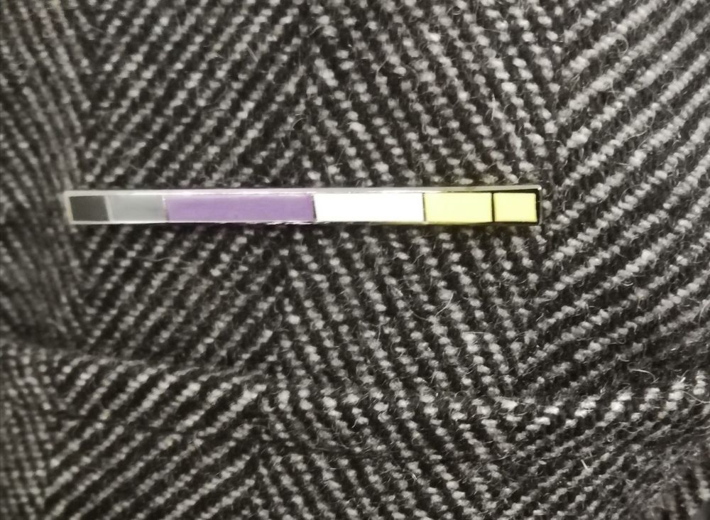A badge pinned on grey fabric. The badge is a narrow bar, divided into segments of irregular size. Each segment has a different color, from left to right the colors are: black, dark gray, purple white, pale yellow, yellow