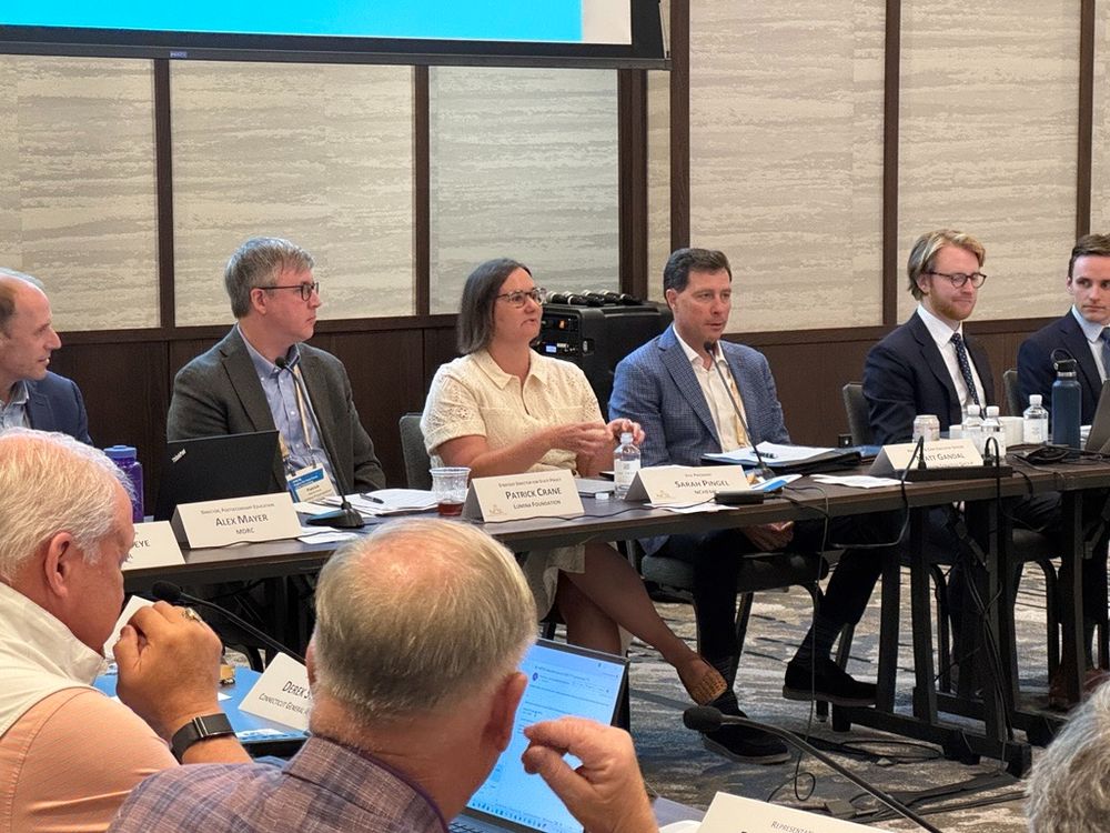 Photo of a conference room with several people, a mix of older and younger adults, sitting at a long, U-shaped table. The focus of the photo is NCHEMS Vice President Sarah Pingel, who is talking on a panel with a large display screen behind her. 