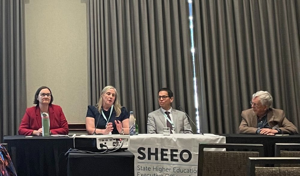 Photo of four people sitting at a black table in a conference room for a panel. On the table is a banner for SHEEO. 