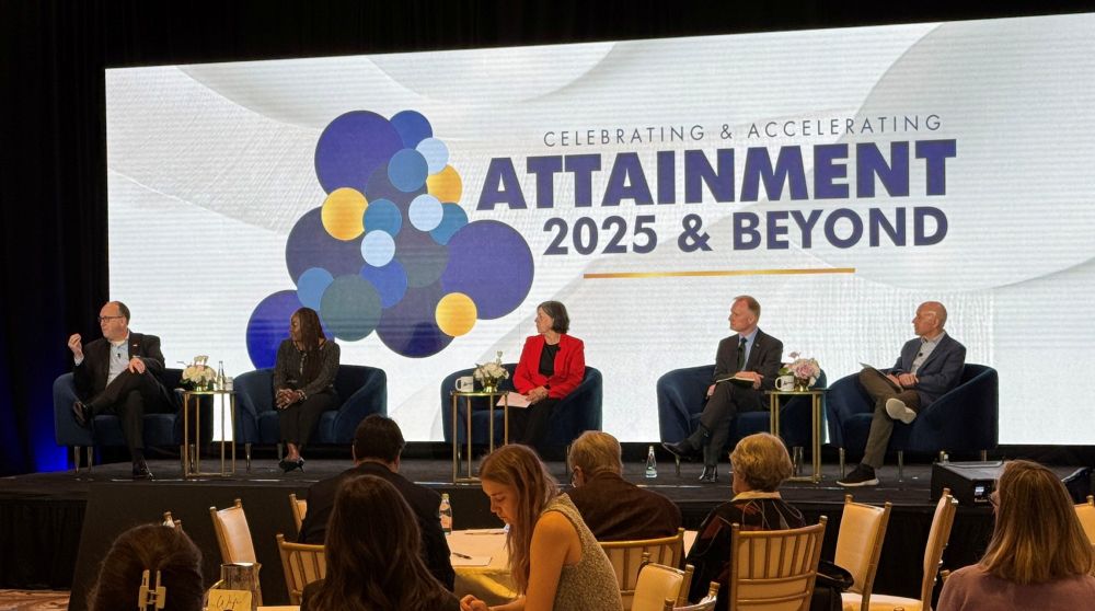 Photo from the Celebrating and Accelerating Attainment 2025 & Beyond conference with a stage where five individuals are seated in a panel discussion. The room has a banner with the conference title and logo behind the stage, with each panelist sitting on a blue chair and attendees visible in the foreground. 