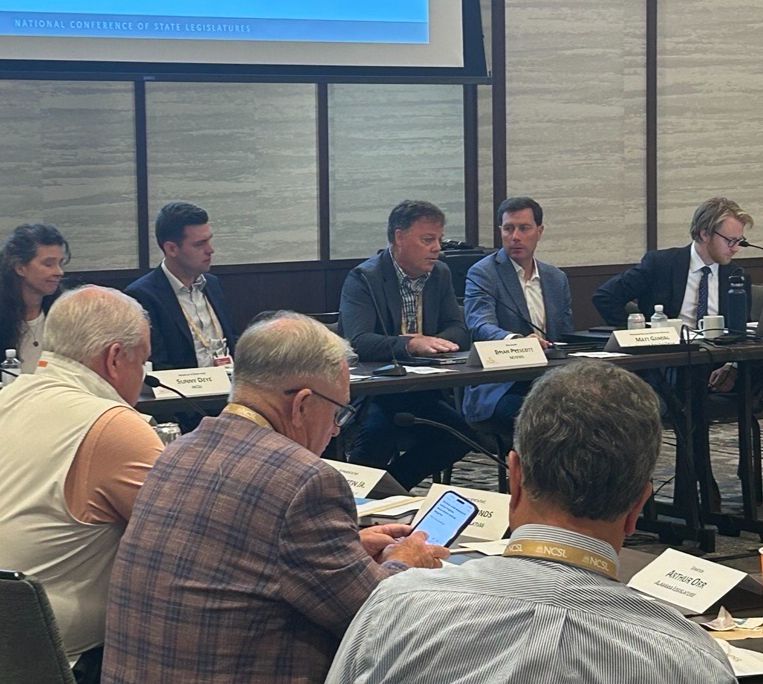 Photo of a conference room with several people, a mix of older and younger adults, sitting at a long, U-shaped table. The focus of the photo is NCHEMS President Brian Prescott, who is talking on a panel with a large display screen behind him. 