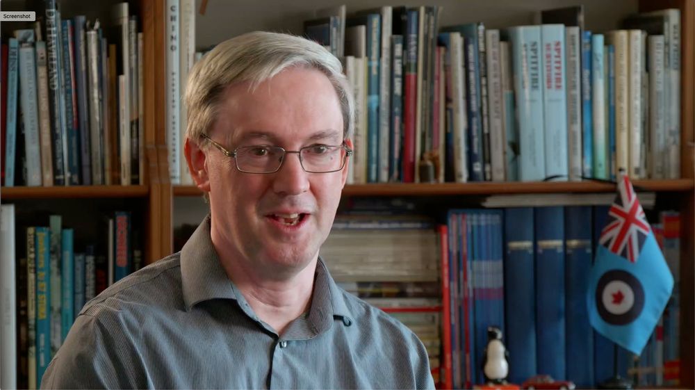 James D Kightly, Aviation Cultures Inc President, talking aviation in front of a bookshelf with a miniature RCAF flag visible among the books.