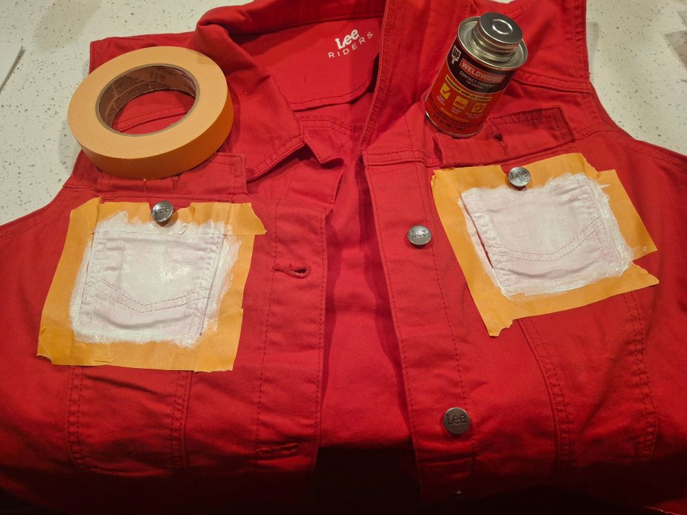 An image of a cropped red denim jacket that's sleeveless. There are 2 squares taped off on the individual breast pockets that have been primed for fabric painting later. 