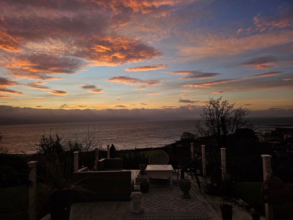 A view from my doorstep over the Firth of Clyde. Pink clouds are scattered across a blue and orange sky. The whole scene glows a warm pinkish, orange hue.