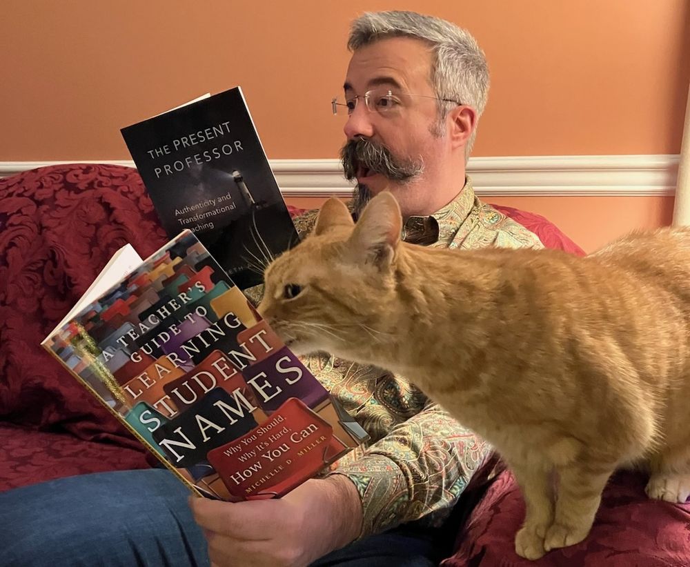 Tom Tobin sits on the couch, reading *The Present Professor* by Liz Norell, while his orange cat Webster sits on his leg, appearing to read *Learning Student Names* by Michelle Miller.