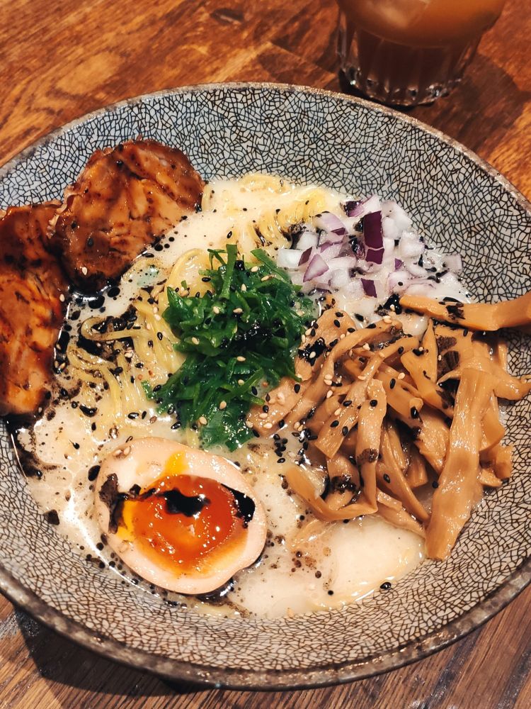 A bowl of Japanese noodle soup, Ramen, on top of a brown wooden table. It's a creamy broth with ramen noodles, chicken slices on the left, a marinated egg half, a good portion of menma slices on the right, some red onions above and spring onions in the middle with sesame sprinkled on top.
