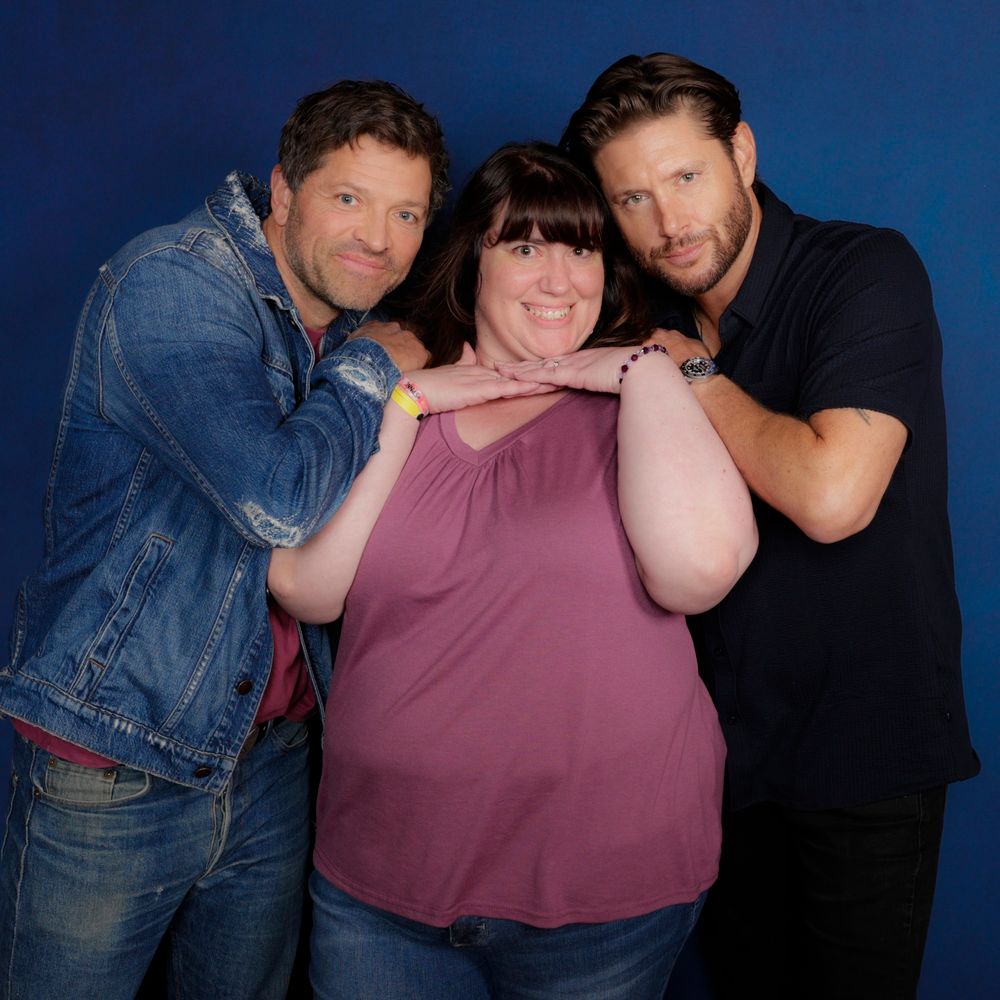 Misha Collins wearing a distressed blue denim jacket with jeans, Jensen Ackles wearing a black short sleeved shirt and black pants. Both are leaned in close to me with hands on my shoulders. I'm in the middle wearing jeans and a rose colored shirt smiling.